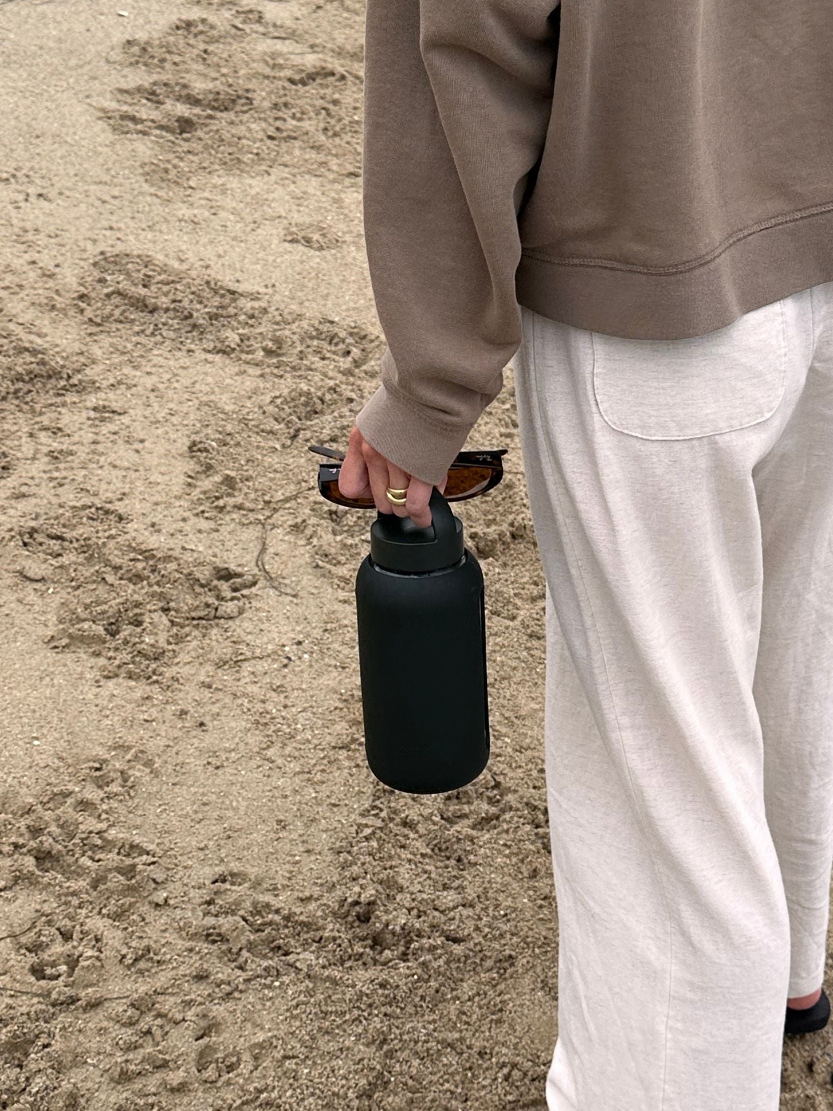 Woman holding the Saltyface x Bink The Day Bottle in one hand with sunglasses walking on the beach. 