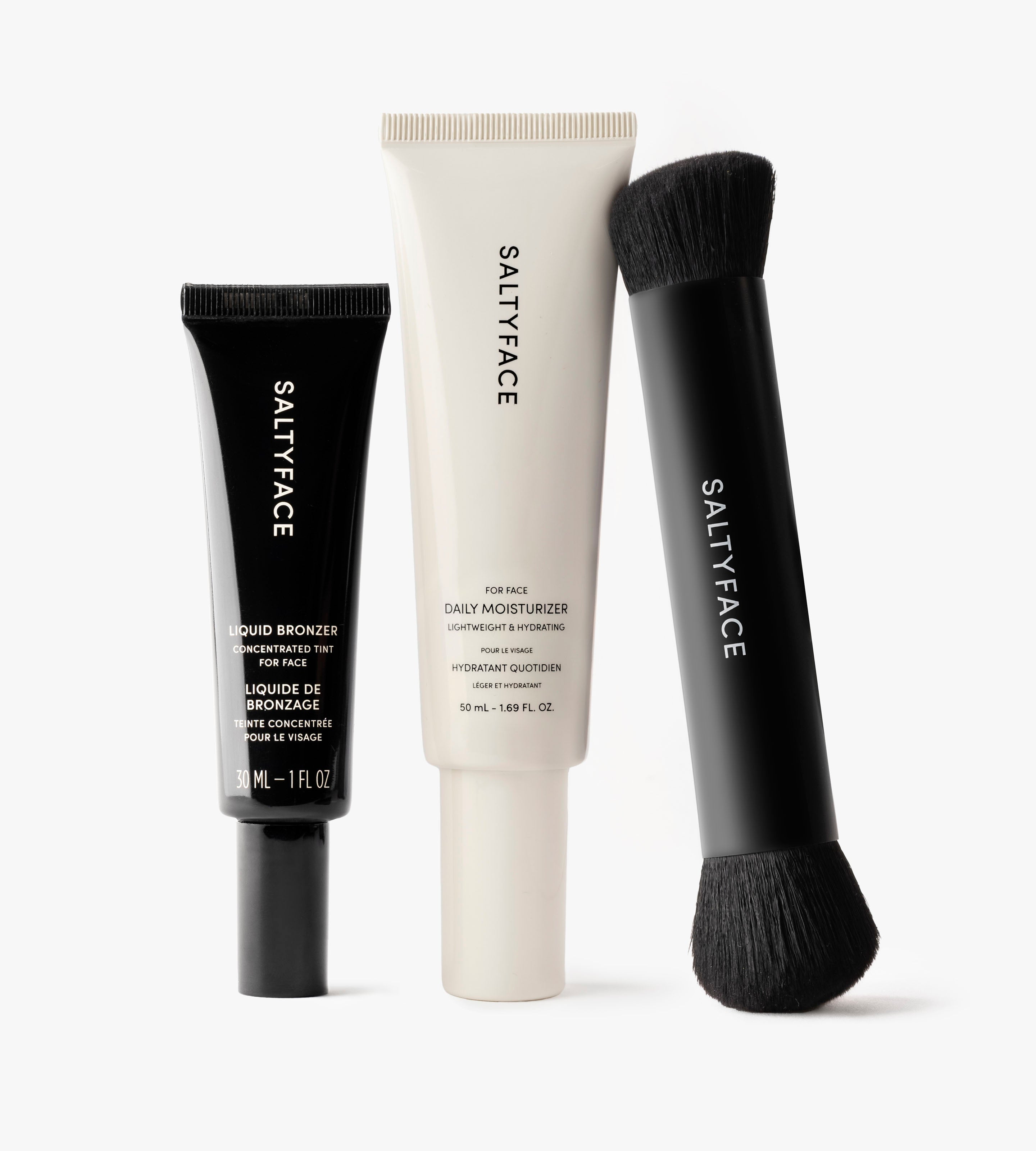The Hydrate & Bronze Set by SALTYFACE includes a black Liquid Bronzer tube, a white Daily Moisturizer tube, and a black dual-ended makeup brush, all upright against a white background.